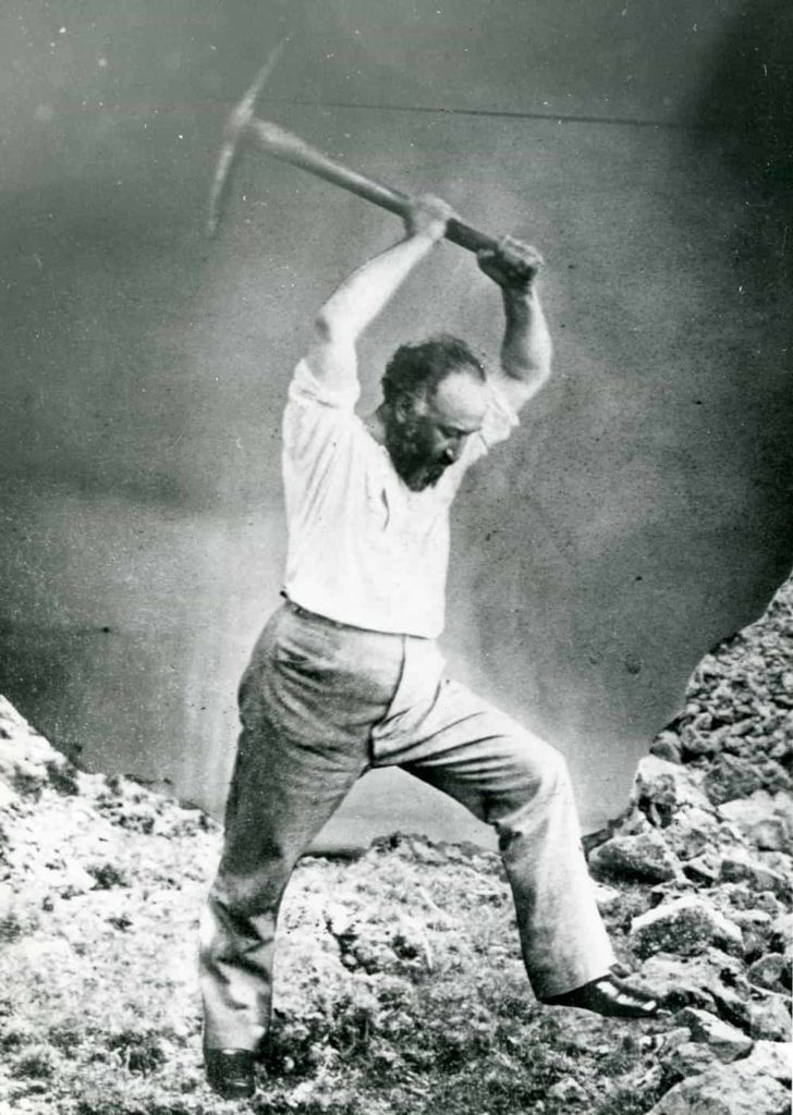 Black and white photograph of Adolph Sutro raising a pick axe over his head to strike rocks