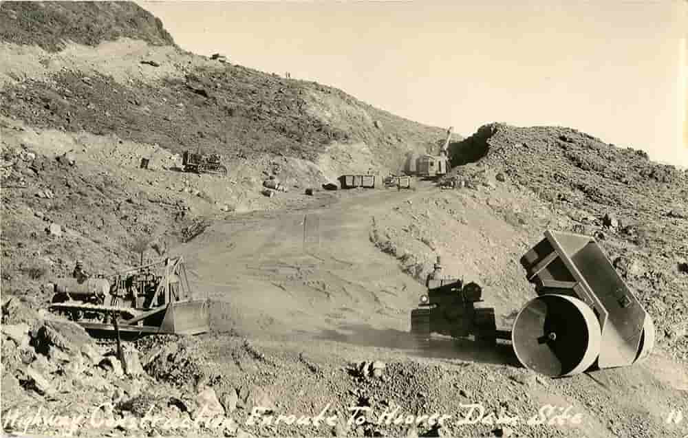Black and white photograph of road construction, construction equipment around a graded dirt road going over a hill.